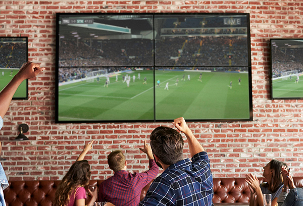 Customers at a restaurant cheering watching soccer on a tv screens.